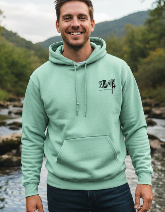 Man wearing a light green hoodie with a logo, standing outdoors by a river with mountains in the background.