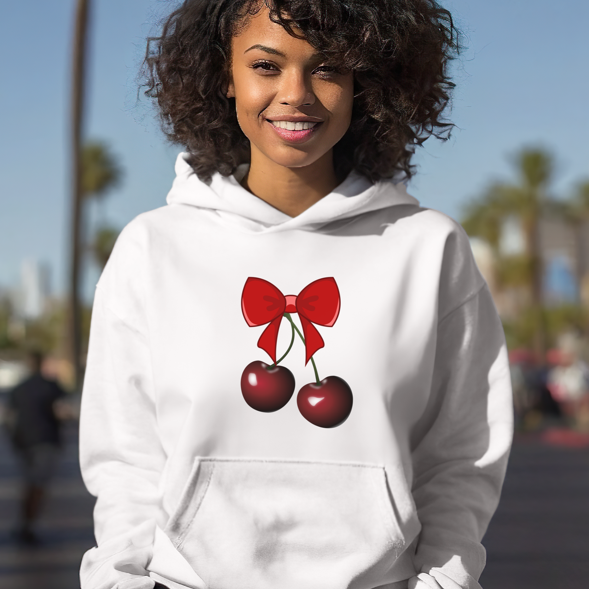 Person wearing a white hoodie with a red bow and cherries design outdoors.